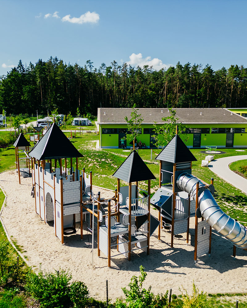 Full view of a large inclusive playground at a camping site. The playground is castle themed, and in the background you can see camper vans.